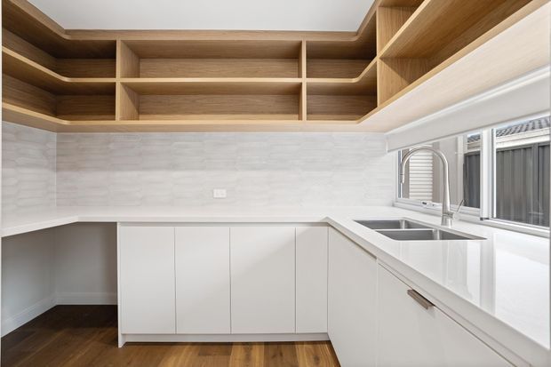 Bright contemporary kitchen featuring white handleless base cabinetry, a white engineered-stone benchtop, stainless-steel double sink and gooseneck tap, and a light timber open-shelving system wrapping across the upper walls. The backsplash is finished in pale textured tile, with timber flooring and ample natural light from the windows.