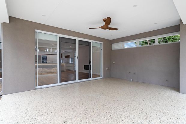 A bright enclosed outdoor living area or patio with a light terrazzo-style floor, textured taupe walls, white ceiling, ceiling fan, high horizontal louvred windows, and large sliding glass doors opening to a modern interior. The space is clean and minimally furnished, with the kitchen and living areas visible beyond the glazing.