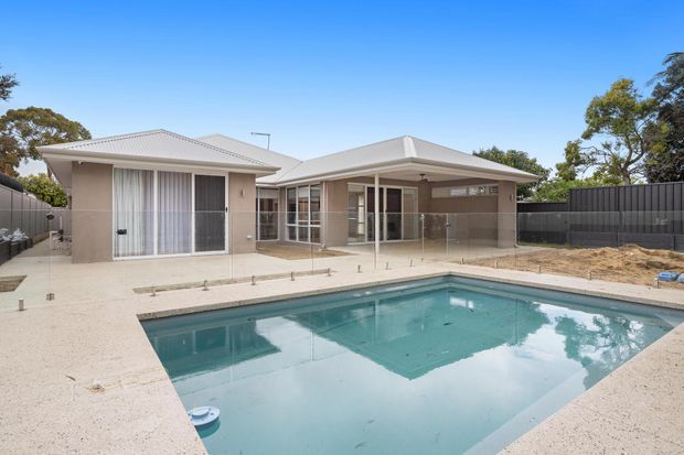 Single-storey contemporary residence viewed from the rear, with pale rendered walls, low-pitched light metal roofs, extensive glazing, a covered outdoor entertaining area, and a rectangular swimming pool surrounded by a light concrete terrace. Dark privacy fencing and established trees frame the property.