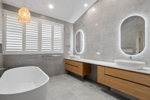 Contemporary bathroom with a freestanding white bathtub, twin white vessel basins, floating timber vanity cabinetry, large grey stone-look wall and floor surfaces, plantation shutters, and rounded-rectangle mirrors. Recessed ceiling lighting and a warm woven pendant create a clean, refined atmosphere.