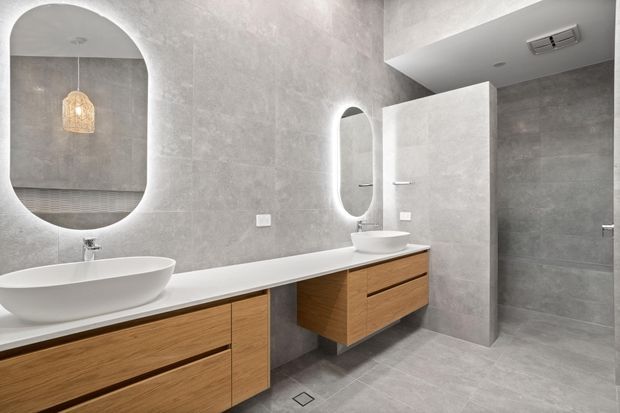 Contemporary bathroom with full-height grey stone-look wall and floor tiles, warm timber floating vanity cabinetry, white vessel basins, an integrated bathtub, rounded backlit mirrors, and a partially screened walk-in shower. The space has a clean, refined, spa-like appearance.