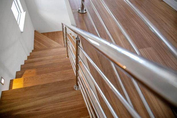 Interior view looking down a modern staircase with warm timber flooring and matching timber-clad wall surfaces. The stair is enclosed by polished horizontal metal balustrades with slender vertical supports, while white walls and a small high-set window provide contrast. The image focuses on the stairway circulation space and contemporary material detailing.