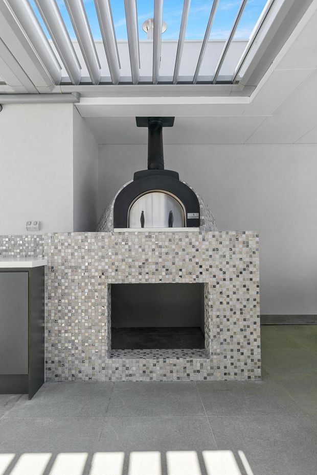 A bright contemporary interior centered on a built-in dome-shaped pizza oven with a black flue and arched opening. The oven surround is clad in small multicoloured mosaic tiles, with a large open firewood storage niche beneath. A skylight or glazed roof opening with parallel structural fins casts strong linear shadows across the grey tiled floor. Partial grey cabinetry is visible at left.