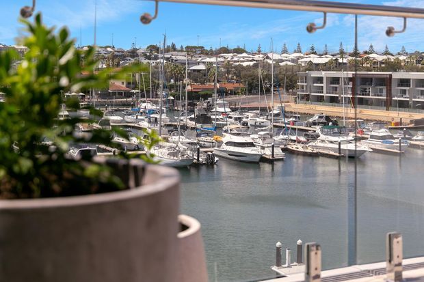 View from a balcony or terrace overlooking a marina with numerous boats, calm water, and waterfront apartment buildings. Large potted greenery and overhead balcony fixtures frame the foreground, with suburban housing and trees visible across the harbour.