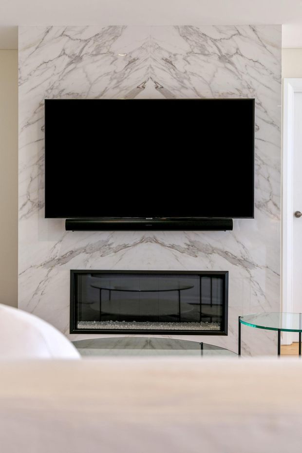 Close-up interior view of a contemporary living room feature wall clad in bookmatched white and grey-veined marble. A wall-mounted television and soundbar sit above a sleek black linear fireplace with glass front and stone-filled base. A curved glass side table is visible at right, with soft neutral furnishings in the foreground.