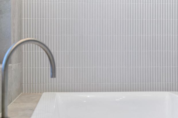 Close-up of a minimalist bathroom with a white freestanding bathtub, curved brushed-metal wall-mounted faucet, pale stone-look surfaces, and vertically ribbed light-grey wall tiles. The composition is clean, bright, and highly textural.