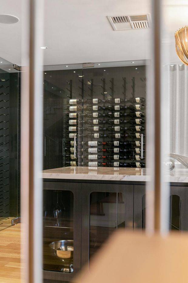 A refined residential wine storage area viewed through vertical foreground elements, featuring a glass-enclosed wine display with horizontally arranged bottles, dark timber cabinetry, a pale stone benchtop, mirrored or glazed surfaces, and integrated lighting. Part of a wine room or home bar is visible, with a warm, sophisticated finish palette.