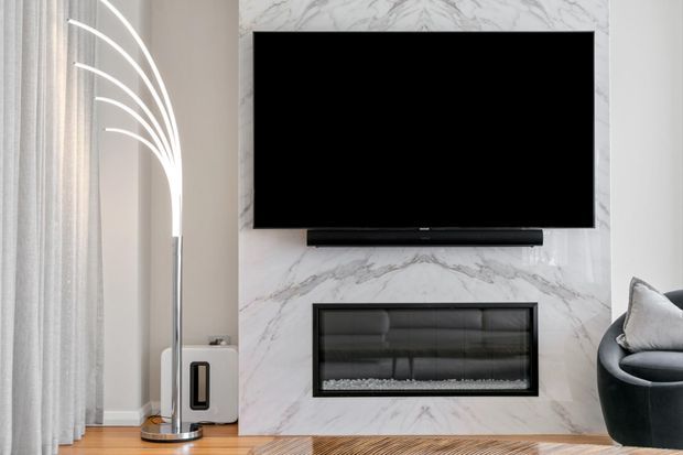 A refined living room media wall featuring a large wall-mounted television, soundbar, and a linear built-in fireplace set within a dramatic white-and-grey veined marble-look feature wall. A sculptural arched floor lamp, sheer curtains, timber flooring, and a dark curved lounge chair complete the space.