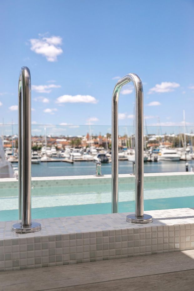 A bright outdoor pool area overlooking a marina. The foreground shows a tiled pool edge with polished stainless-steel entry rails, turquoise pool water, and a glass balustrade. Boats and waterfront buildings are visible beyond the pool under a clear blue sky.