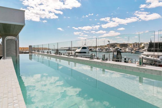 A rooftop or elevated infinity-edge swimming pool overlooking a marina, with boats, waterfront residences, and a bright blue sky beyond frameless glass balustrades. The pool is lined in pale aqua tiles and bordered by light paving, creating a contemporary resort-like outdoor setting.