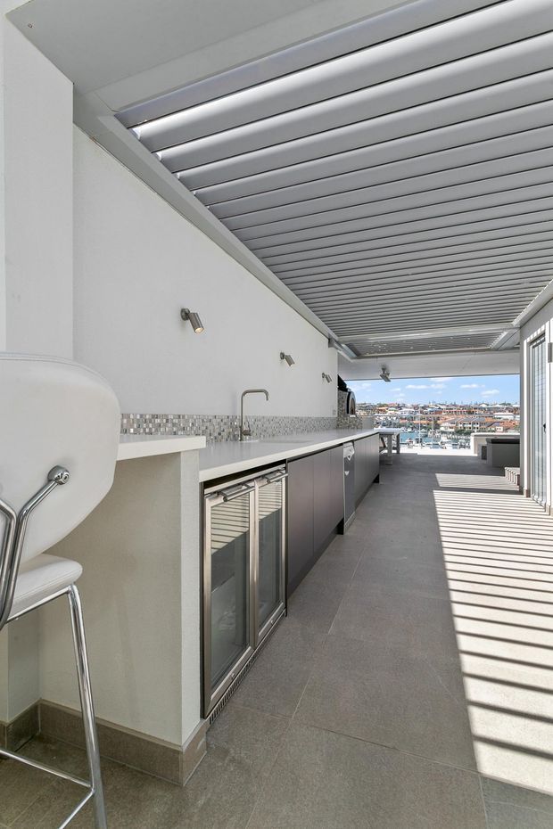 A bright, contemporary outdoor entertaining area with a long built-in bar kitchen, stainless-steel refrigeration, sink, tiled benchtop and mosaic-tile splashback. A louvred pergola roof creates strong linear shadows across the grey tiled floor, with marina and urban views visible at the far end.