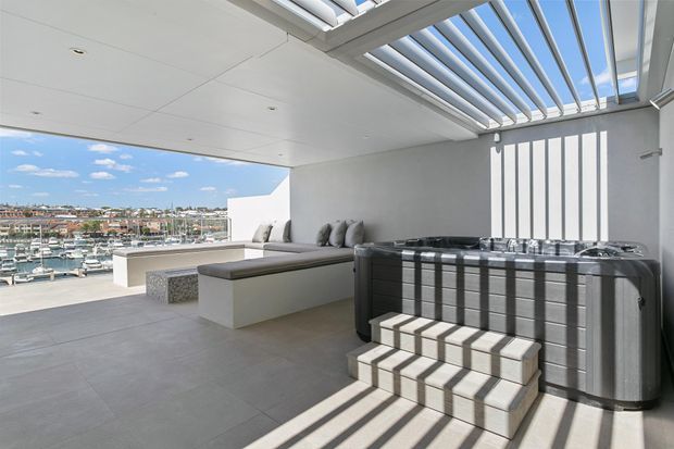 A bright, contemporary rooftop terrace or balcony with expansive views over a marina and surrounding urban landscape. The covered space features a built-in pale grey sectional bench, a low rectangular table, large-format grey floor tiles, and a freestanding spa positioned beneath a slatted retractable roof or pergola. Strong linear sunlight and shadows create a crisp architectural effect.
