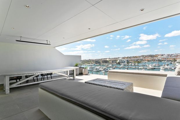 A bright, expansive covered outdoor terrace or rooftop lounge overlooking a marina and waterfront buildings. The space features a long white dining table with benches, built-in upholstered seating, a low patterned coffee table, pale tiled flooring, a white ceiling with recessed lighting, and wide open glazing or balcony openings framing the harbour view.