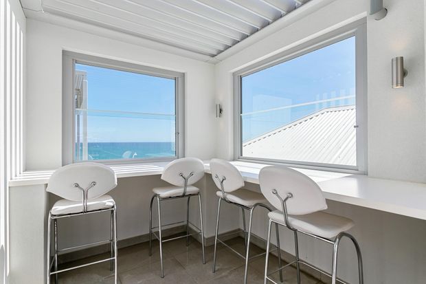 Bright coastal breakfast bar or casual dining nook with four white bar stools arranged along a built-in white counter beneath large corner windows. The windows overlook blue ocean water and a neighbouring light metal roof. White ribbed ceiling, white walls and trim, grey tiled flooring, and simple contemporary finishes create an airy, clean interior.