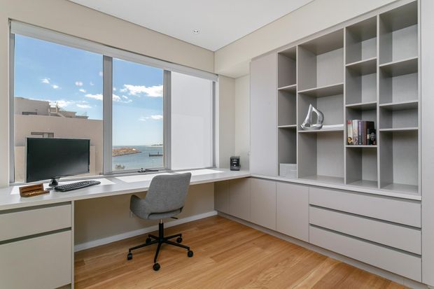 A bright, contemporary home office with a built-in desk running beneath large windows and a wall of bespoke open shelving and closed drawers. The room has pale neutral walls and joinery, warm timber flooring, a grey upholstered swivel chair, and views toward the water through the glazing.
