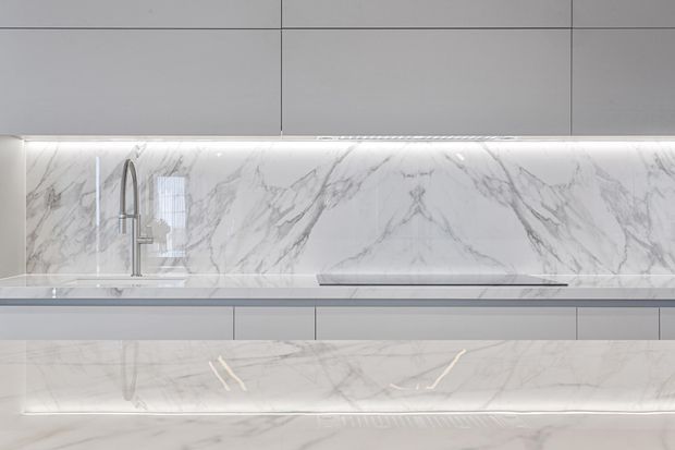 Close-up of a sleek contemporary kitchen featuring grey flat-panel cabinetry, a marble-look illuminated splashback, a matching stone benchtop with integrated cooktop, and a tall brushed-metal mixer tap. Under-cabinet lighting highlights the veined stone surfaces and minimalist joinery.