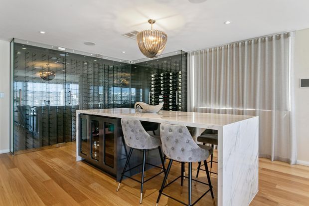 A refined residential kitchen and entertaining space featuring a large white marble-look island with waterfall ends, upholstered bar stools, warm timber flooring, and a dramatic glass-enclosed wine cellar with mirrored shelving. A sculptural pendant light and full-height curtains add a polished, luxurious feel.