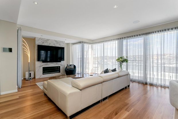 Bright contemporary living room with a large cream sectional sofa, timber flooring, marble-look fireplace and TV feature wall, and expansive floor-to-ceiling windows dressed with sheer curtains. The room has a clean, open layout with city views beyond the glazing.