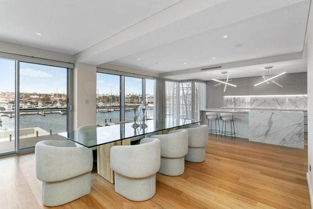 Bright contemporary open-plan dining and kitchen interior with floor-to-ceiling glazing overlooking a marina. The room features warm timber flooring, a large glass dining table with rounded upholstered chairs, a white marble kitchen island and backsplash, streamlined cabinetry, and sculptural linear pendant lighting.