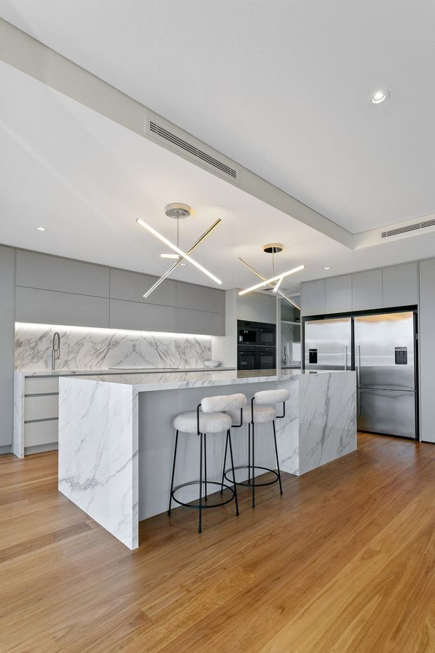A bright, contemporary kitchen with a large white marble-look island and waterfall ends, matching marble-look splashback and benchtops, matte grey full-height cabinetry, integrated dark appliances, and a stainless-steel refrigerator. Two upholstered bar stools sit at the island. Warm timber flooring contrasts with the cool grey and white finishes, while slim linear pendant lights and recessed ceiling lighting illuminate the space.