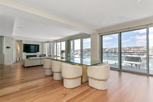 Bright, open-plan waterfront apartment interior with a living room and dining area overlooking a marina through expansive floor-to-ceiling glazing. The space features warm timber flooring, white walls and ceilings, a glass-topped dining table with rounded upholstered chairs, and a neutral sectional sofa with a wall-mounted television. The overall design is clean, contemporary, and airy.
