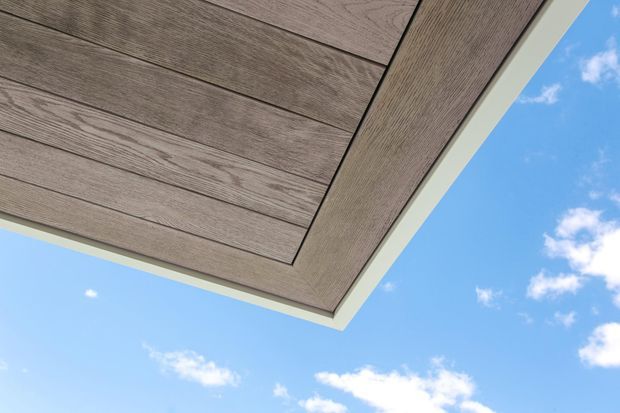 Close-up upward view of a contemporary exterior soffit or overhang clad in wide grey-brown wood-look boards, with a contrasting pale trim edge against a bright blue sky. The image focuses on the building’s underside and clean linear detailing.