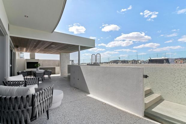 A contemporary rooftop terrace with an outdoor lounge and dining area beneath a timber-lined covered canopy. The terrace features textured grey flooring, a large woven sectional sofa, dining chairs, a wall-mounted outdoor television, and a partially enclosed swimming pool accessed by steps. The scene has expansive coastal or urban views under a bright blue sky.