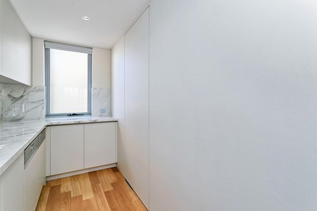 Compact contemporary galley kitchen with white flat-panel cabinetry, integrated appliances, a pale marble-look splashback and benchtop, a single window, and warm timber flooring. The space is bright and minimalist, with full-height white storage on the right wall.