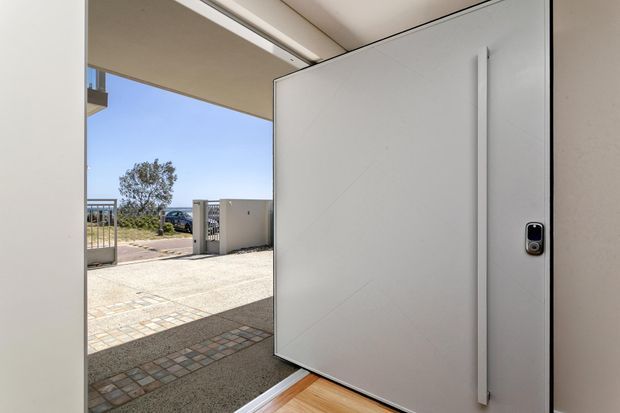 View from inside a garage through a large open sectional garage door toward a paved driveway and front gate. The scene features a clean white door and walls, a timber-look interior floor threshold, and a bright suburban exterior.
