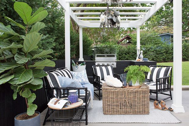 A covered outdoor entertaining area beneath a white timber pergola, furnished with striped lounge chairs, a wicker coffee table, cushions, and a barbecue kitchen. Dark privacy screening, a striped outdoor rug, potted greenery, and surrounding garden vegetation create a relaxed garden setting.