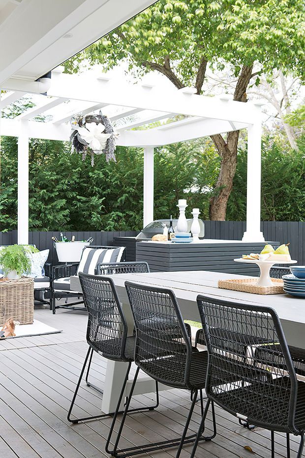 A bright covered outdoor entertaining area with a large dining table, black woven dining chairs, a secondary seating area, and a built-in dark outdoor bar or kitchen counter. White pergola beams and a translucent roof frame mature greenery surrounding the timber deck.