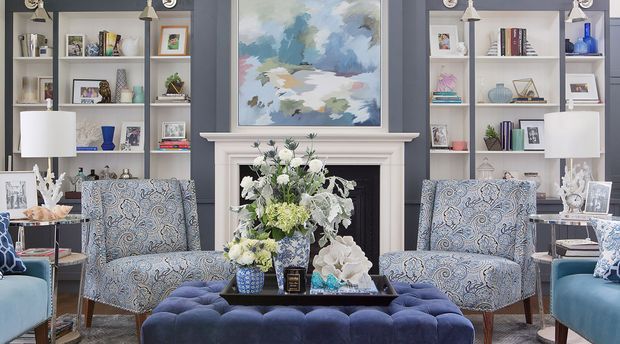 A refined, symmetrical living room with deep blue-grey walls, built-in white shelving flanking a white fireplace, and a large abstract landscape artwork above the mantel. Two patterned armchairs face a tufted navy ottoman and central floral arrangement, with table lamps, books, decorative objects, and blue upholstered seating completing the room.
