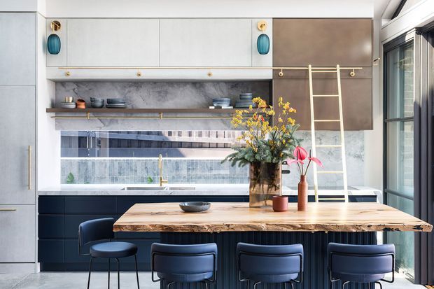 A sophisticated contemporary kitchen with a large timber island, dark ribbed cabinetry, integrated seating, open shelving, and a decorative ladder rail. Pale upper cabinetry, metallic accents, patterned backsplash glazing, and abundant daylight create a refined, high-contrast interior.