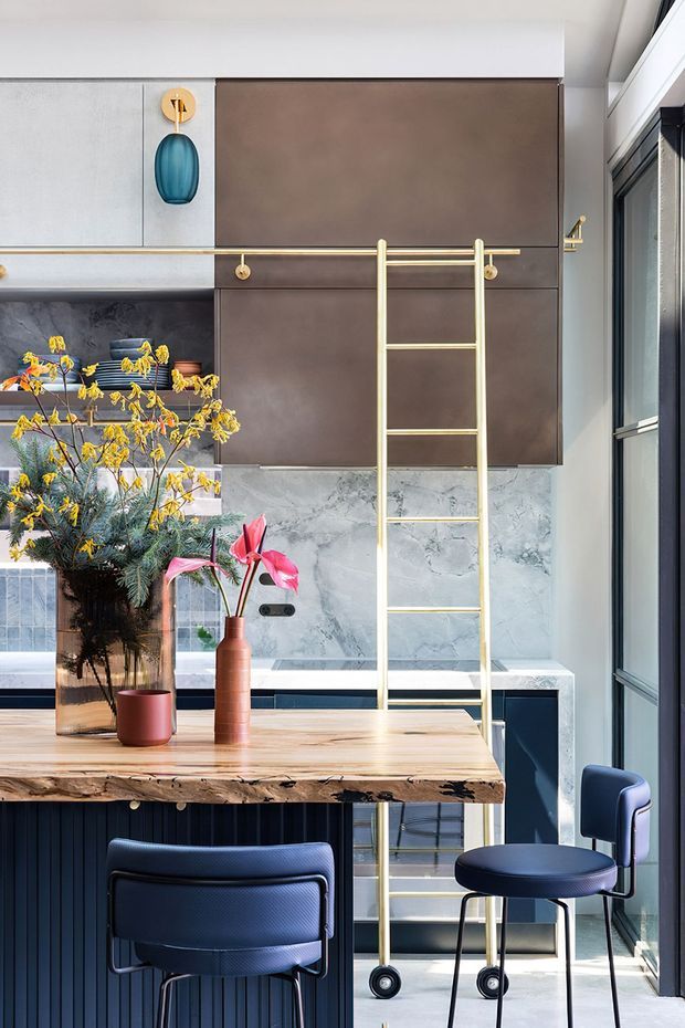 A refined contemporary kitchen and dining area with dark brown upper cabinetry, a veined grey marble splashback, pale timber dining table, dark fluted cabinetry, and brass detailing. A decorative brass ladder leans against the cabinetry, while navy upholstered dining chairs, colourful floral arrangements, and large black-framed windows add visual contrast. The compact space is bright and vertically oriented, with a strong mix of timber, marble, dark cabinetry, and warm metallic accents.