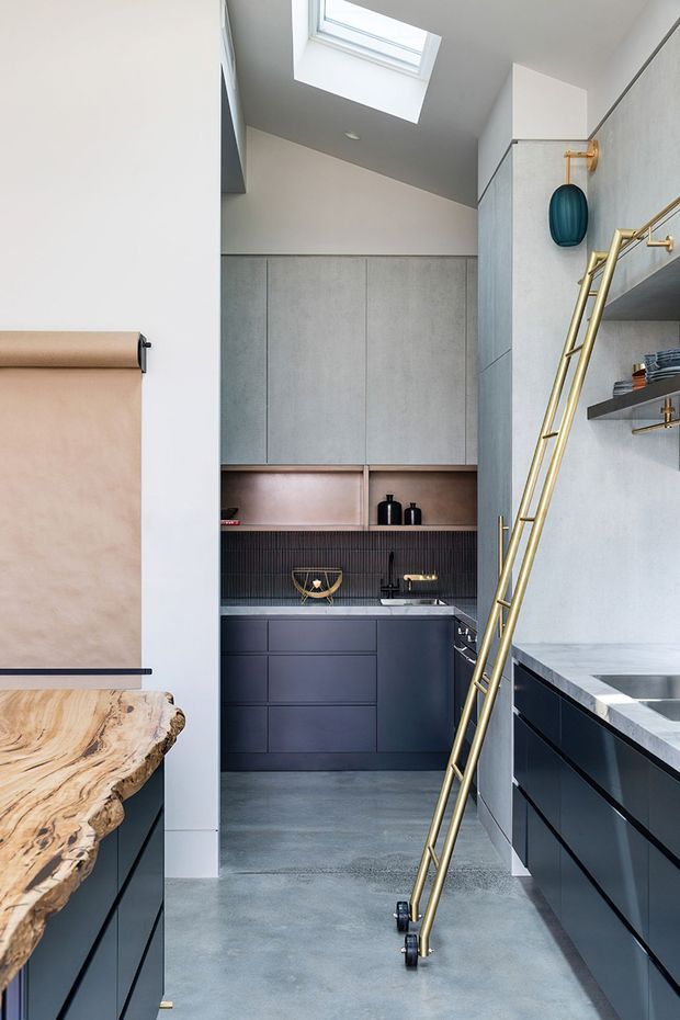 A narrow, high-ceilinged contemporary kitchen with dark blue-grey cabinetry, pale concrete-look wall panels, a dark vertically ribbed splashback, and a grey polished concrete floor. A skylight brings natural light into the space. Brass shelving and a leaning brass ladder add a strong metallic accent, while a sculptural timber edge is visible in the foreground.