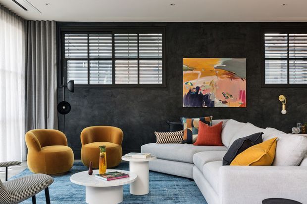 A contemporary living room with a large pale sectional sofa, mustard and rust accent cushions, sculptural white nesting coffee tables, and mustard velvet lounge chairs. The room features a dark textured feature wall, black-framed horizontal windows with timber shutters, full-height curtains, a blue area rug, and colourful abstract artwork.