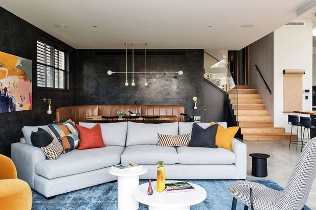 A contemporary open-plan living room with a large pale sectional sofa, colourful cushions, round white coffee tables, and a blue rug. The room features a dark textured feature wall, integrated leather banquette seating, black-framed louvred windows, statement pendant lighting, and a timber staircase with glass balustrade leading to an upper level.