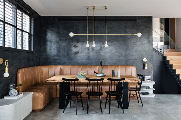 A sophisticated open-plan dining and living area with a long brown leather banquette wrapping around a dining table, surrounded by black spindle-back chairs. The room features a dark textured feature wall, polished concrete flooring, black-framed louvred windows, sculptural brass-and-globe pendant lighting, and a staircase with timber treads and glass balustrading.