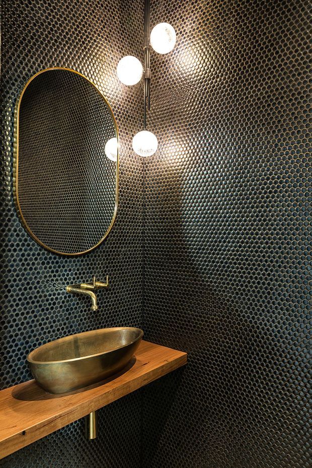 A dramatic compact bathroom or powder room clad entirely in small dark circular mosaic tiles. The space features a floating natural timber vanity shelf, a brushed brass vessel basin and wall-mounted tapware, an oversized rounded-rectangle mirror with a brass frame, and a vertical cluster of spherical wall lights. The dark reflective tile surface and warm metallic and timber accents create a luxurious, moody atmosphere.
