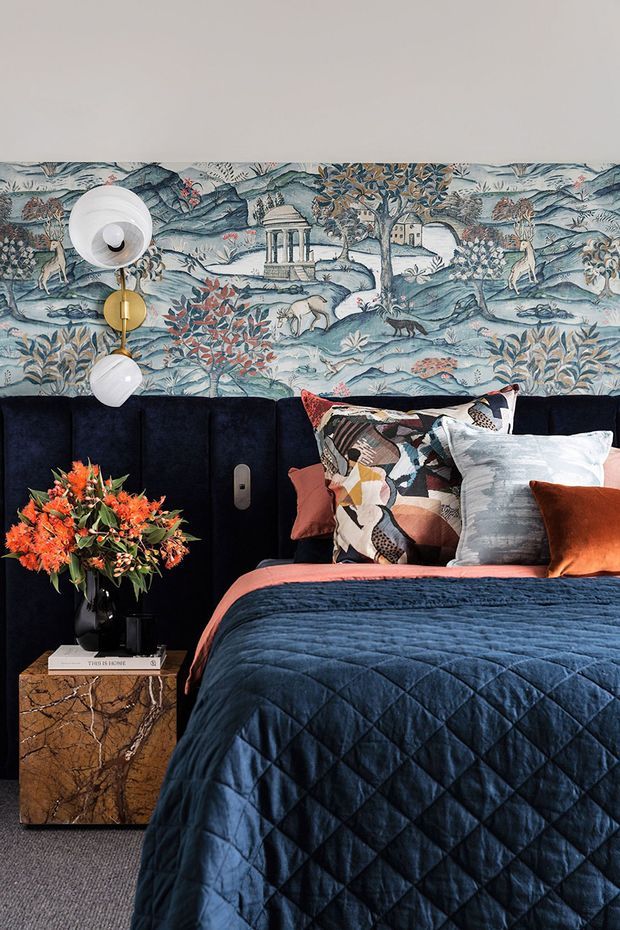 A richly styled bedroom vignette featuring a deep navy upholstered headboard, navy quilted bedding, layered patterned and solid cushions, and a vibrant floral arrangement on a dark marble-patterned bedside table. The wall above the headboard is covered with a scenic blue-toned botanical and landscape wallpaper, complemented by a sculptural brass-and-white wall light. The room has a dramatic, eclectic and luxurious character with warm orange and terracotta accents.
