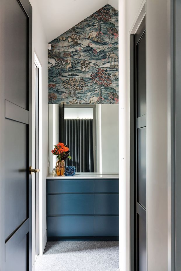 A narrow, refined hallway or dressing-area view leading to a compact vanity with deep blue built-in drawers and a pale stone benchtop. The space features dark framed-panel doors, a large mirror, charcoal curtains, grey carpet, and a striking botanical toile wallpaper extending up the triangular ceiling wall. A vase of orange flowers adds colour to the otherwise cool, sophisticated scheme.