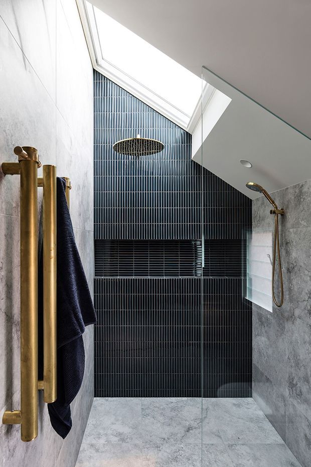 A contemporary walk-in shower bathroom with a frameless glass screen, large skylight, dark narrow vertical wall tiles, and pale grey stone-look wall and floor tiles. Brass fixtures include a rain shower, handheld shower, and heated towel rail, creating a refined high-contrast scheme.