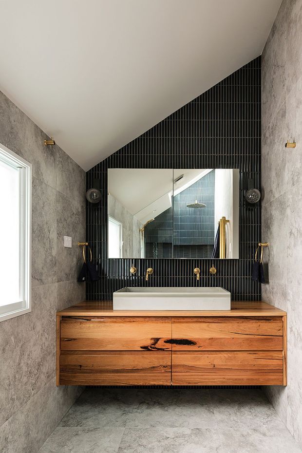 A refined contemporary bathroom featuring a floating natural timber vanity with prominent grain and live-edge character, a white vessel basin, wall-mounted brass fixtures, and a large frameless mirror. The vanity wall is finished in narrow black vertical tiles, contrasting with pale grey stone-look wall and floor finishes. A sloped ceiling creates a distinctive angular form, with wall sconces flanking the mirror.
