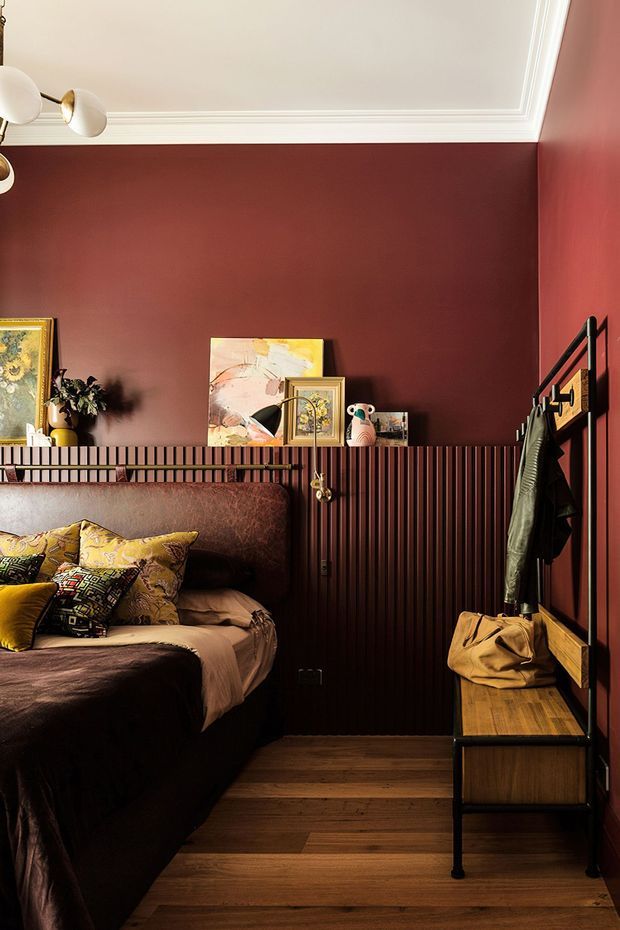 A compact, richly coloured bedroom with deep burgundy painted walls, dark timber flooring, and a half-height vertical timber-panelled wall behind the bed. The room contains a double bed with patterned cushions and dark bedding, framed artwork and a small plant on the ledge, a wall-mounted coat rack, and a narrow timber bedside bench. White decorative ceiling moulding and a brass-and-white wall light add contrast. The styling is warm, eclectic, and dramatic.