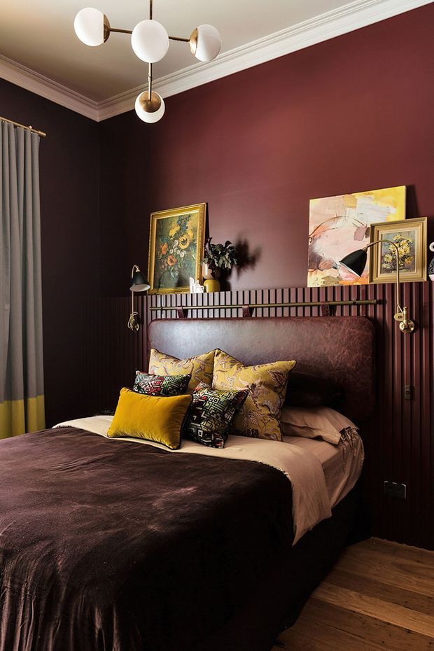 A richly styled bedroom with deep burgundy walls, dark vertical wall panelling, white decorative ceiling moulding, and warm timber flooring. The bed has a tall brown upholstered headboard, dark velvet bedding, layered patterned and mustard cushions, and neutral linen sheets. Framed artwork, plants, wall-mounted reading lamps, and a sculptural globe pendant add an eclectic, moody character.
