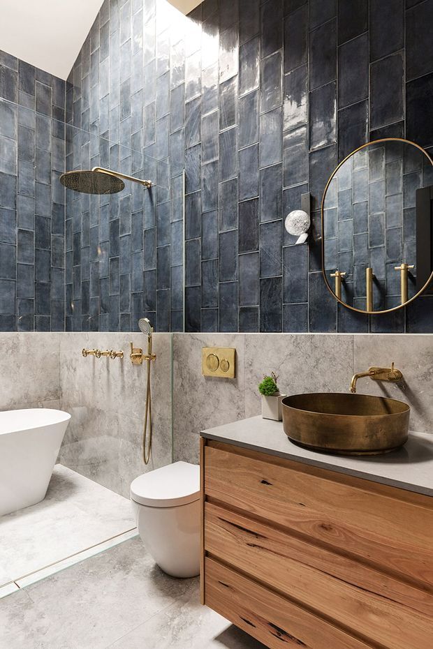 A refined contemporary bathroom featuring a freestanding white bathtub, wall-mounted toilet, walk-in shower, and timber vanity with a dark stone benchtop and bronze vessel basin. The walls combine glossy dark blue-grey rectangular tiles with pale textured stone-look surfaces, complemented by brass/gold tapware, shower fittings, and a large round mirror.