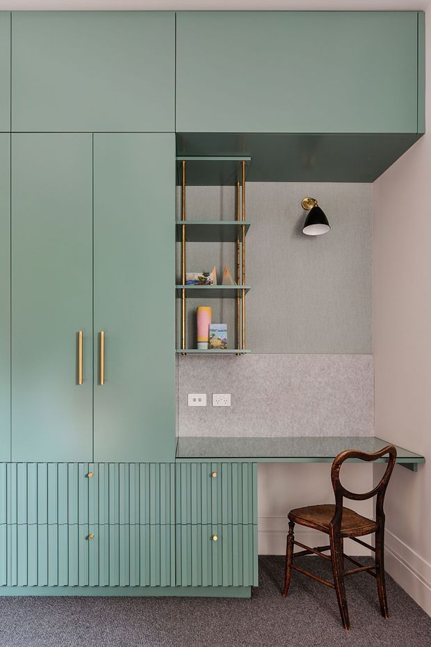 A compact built-in study nook with muted sage-green cabinetry, tall storage doors, a vertically fluted lower cabinet, and brass pulls. Open shelving with brass framing sits beside a textured pale wall panel, beneath a dark projecting overhead cabinet and a small black-and-brass wall light. A vintage wooden chair occupies the desk area, with grey carpet underfoot and pale pink walls.
