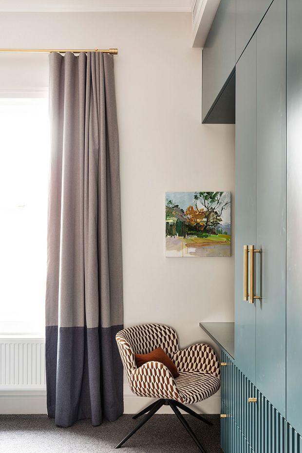A refined interior corner featuring a patterned armchair beside tall two-tone curtains and a large built-in blue-grey cabinet. Brass curtain hardware and cabinet handles add warmth, while a small landscape artwork hangs on the pale wall. The space is softly lit by a large window and finished with carpet flooring and decorative wall and ceiling mouldings.