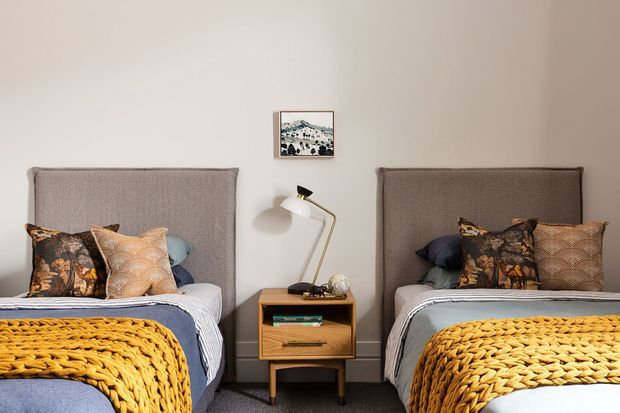 A symmetrical bedroom with two single beds positioned against a warm off-white wall, each featuring a tall upholstered headboard and layered patterned cushions. A small timber bedside table sits between the beds with a slim angled lamp and framed artwork above. Chunky mustard knitted throws add a bold accent over muted blue-grey bedding, while grey carpet covers the floor.
