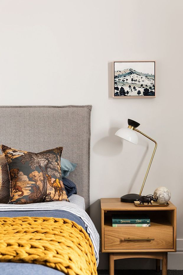 A warmly styled bedroom vignette featuring a textured upholstered headboard, patterned decorative pillows, layered blue and white bedding, and a mustard yellow chunky knit throw. A timber bedside table with drawer storage holds a brass and black articulated lamp, small decorative objects, and books. The pale neutral wall is decorated with a framed landscape artwork.